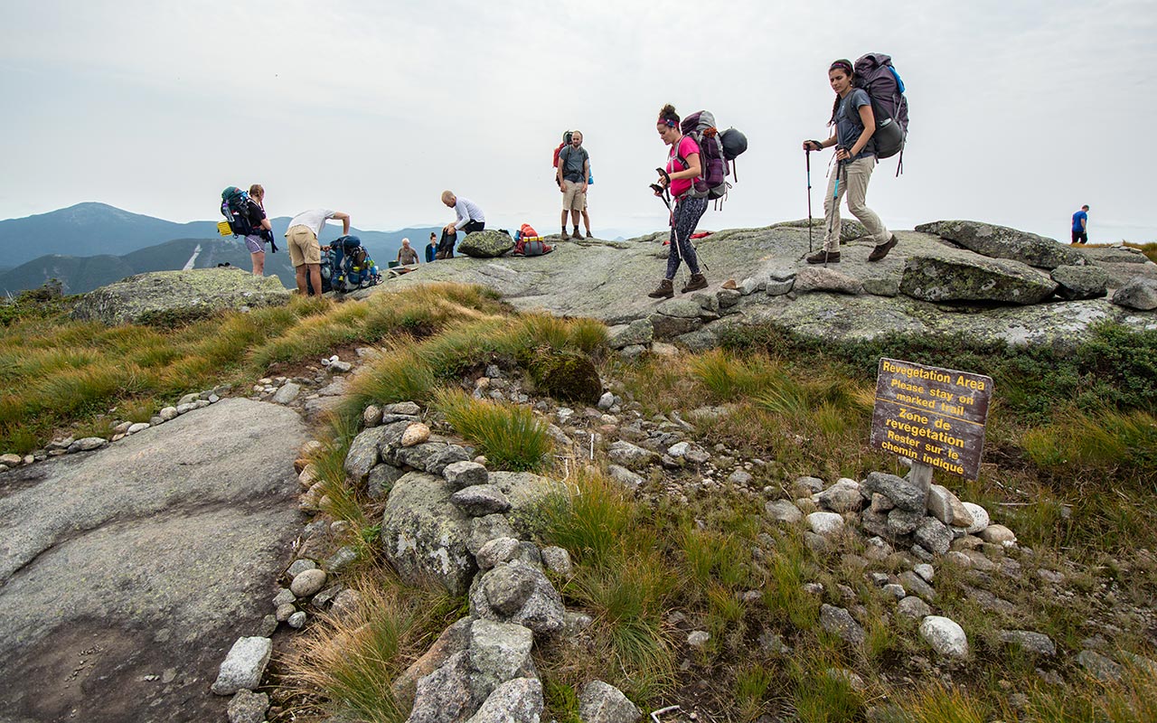 Several people resting or walking atop the rocky mountain summit. A sign warning people to avoid the revegetation area is in the foreground. Photo source: ADK/Seth Jones